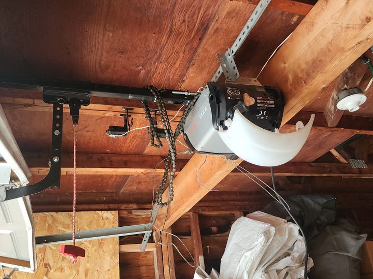 Chain-drive opener fixed to the exposed rafters of an unfinished garage ceiling, with the rail, chain and manual release cord in view